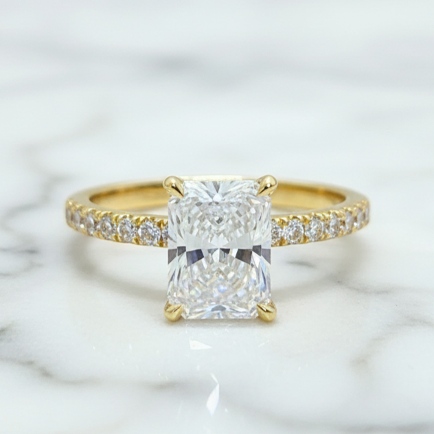 A yellow gold engagement ring featuring a large rectangular radiant-cut diamond center stone, with a pavé band set with small round diamonds, resting on a white marble surface.